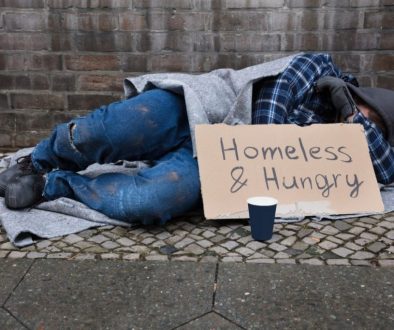 A homeless man lying on the pavement with a "homeless and hungry" sign.