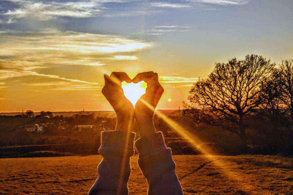 The woman created a heart shape with his hand in front of the sunset.