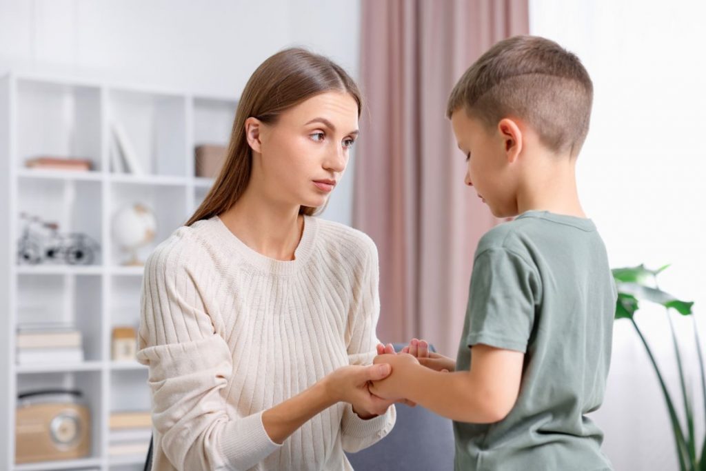 Concerned mother in a cream-colored sweater kneeling to hold her young son's hands, looking directly into his eyes with a gentle but serious expression while he listens, illustrating a moment of guidance in manners, respect, and empathy as elements of social graces.