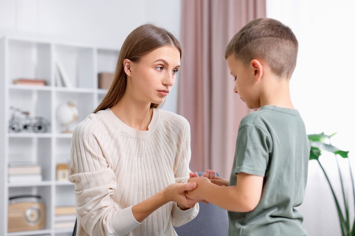 Concerned mother in a cream-colored sweater kneeling to hold her young son's hands, looking directly into his eyes with a gentle but serious expression while he listens, illustrating a moment of guidance in manners, respect, and empathy as elements of social graces.