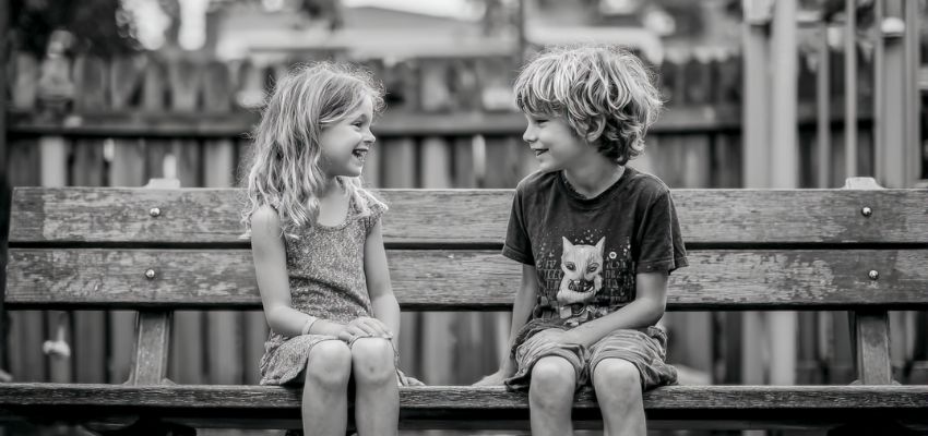 Black-and-white portrait of a smiling young girl and boy sitting together on a wooden park bench, facing each other with friendly expressions and relaxed postures, capturing innocent friendship and polite interaction that reflect early social graces in childhood.