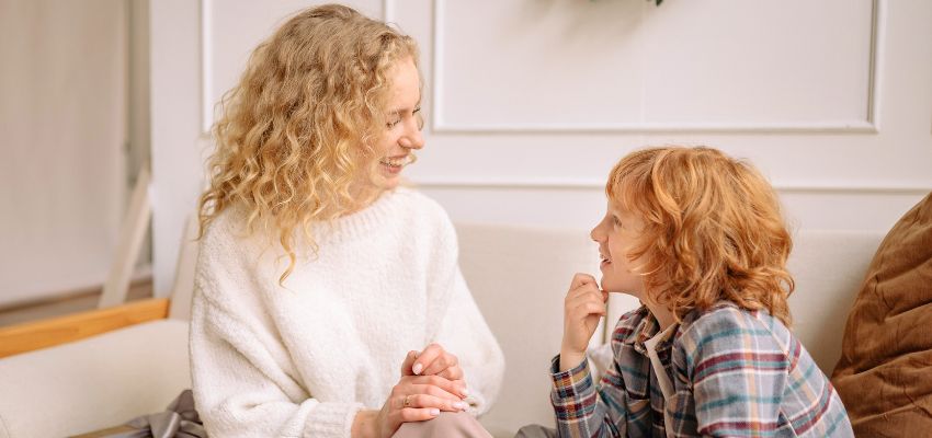 Blonde mother in a cozy white sweater smiling warmly and attentively listening to her young red-haired son as they sit together on a couch, hands clasped, demonstrating kind communication and attentive listening as part of teaching social graces to children.