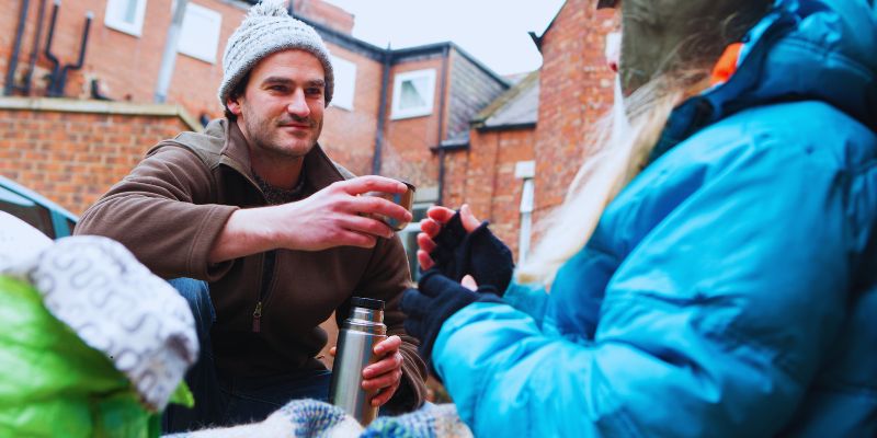 People in cold weather coats gathered outside sharing hot drinks from a thermos near brick buildings, illustrating community support amid hardship and facts about homelessness such as exposure to extreme weather, higher mortality rates nearly 30 years earlier than average, and the human impact of housing instability