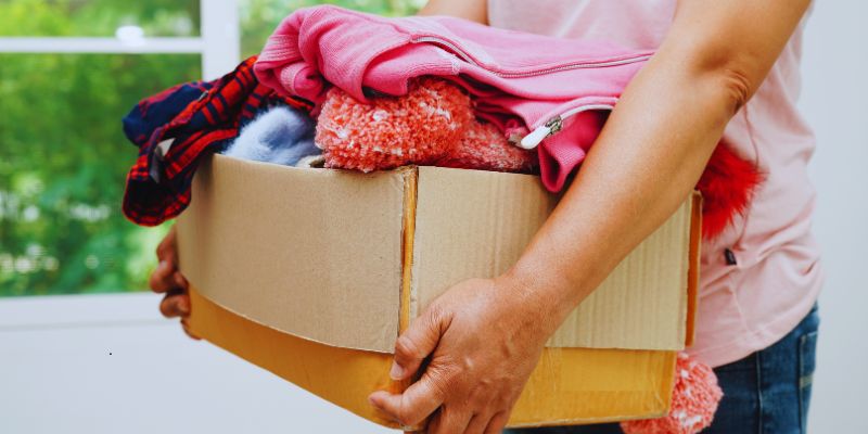 Person holding a large cardboard box filled with folded clothes, sweaters, and cozy blankets, highlighting most needed items for homeless support and donation.