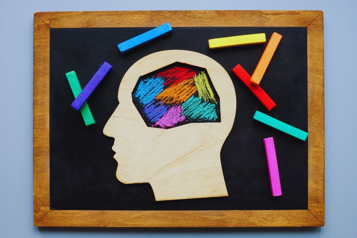 Neurodivergent mind illustrated as a wooden head cutout on a black chalkboard, with a vibrant multicolored brain area drawn in chalk and scattered colorful chalk sticks around it.