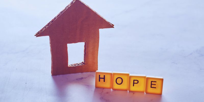 unhoused vs homeless (1) A house made of cardboard with adjacent word blocks forming the word 'hope'.
